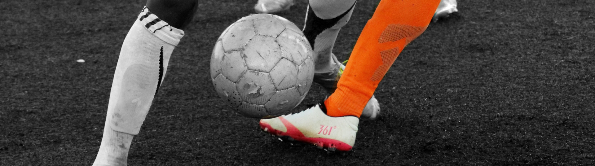 Zwei Füße, einer mit weißen und der andert orangen Stuzen und Schuh, in der Mitte ein Ball
